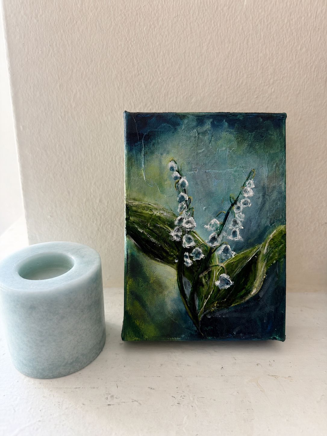 Lilies of the valley soft dark blue background and two stalks of flowers in front of green leaves on a mantle leaning against a neutral colored wall next to a candle