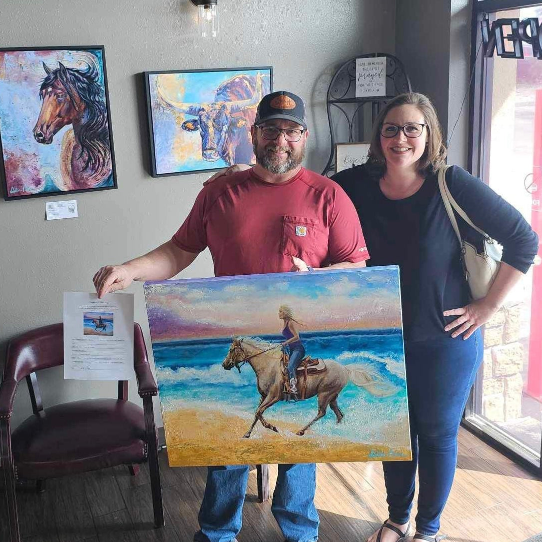 Allison Victoria and man holding his commissioned painting of his wife horseback riding on the beach inside a gallery with other original artworks displayed on the wall