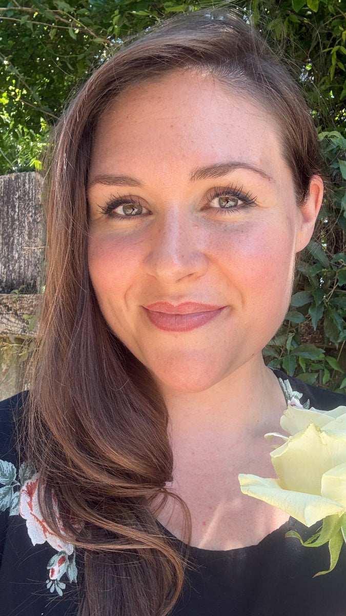 Allison Victoria beautiful woman with green eyes and long brown and sunkissed hair standing in front of a vine covered fence under a tree close up holding a white rose wearing a black dress with some roses on it