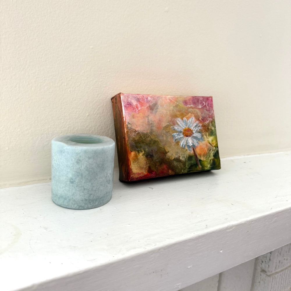 Small painting with a floral design on a wooden frame next to a gray candle holder on a white surface. Side angle.