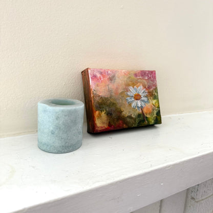 Small painting with a floral design on a wooden frame next to a gray candle holder on a white surface. Side angle.