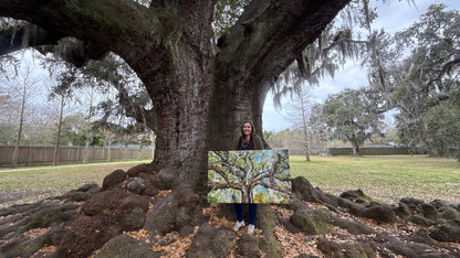 Artist, Allison Victoria, holding a large 30x48 inch painting on gallery wrapped canvas "Beneath the Tree of Life" a large live oak tree, in New Orleans, Louisiana standing outside in Audubon Park, in front of it on the huge roots smiling