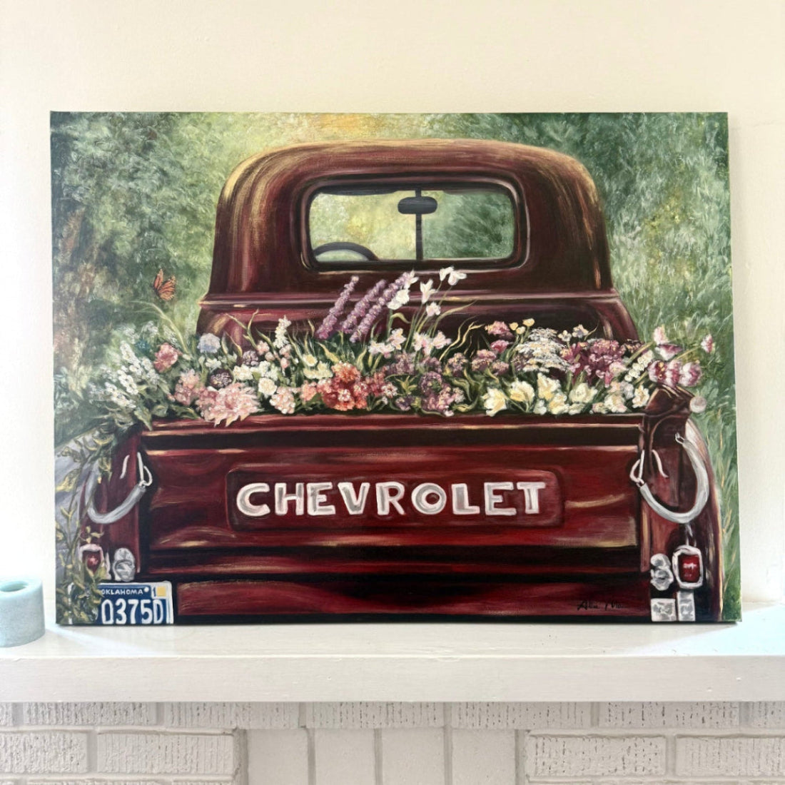 Painting of Old Vintage Red Chevrolet truck with a variety of flowers in the bed on a decorative mantle piece next to a candle, lots of Reds, pinks, greens.