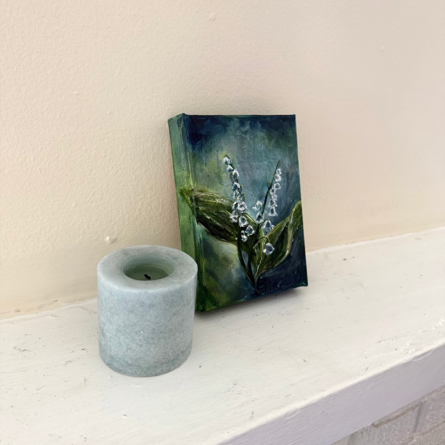Lily of the Valley flower floral acrylic palette knife  floral painting by Allison Victoria deep greens and blues cool tones and texture on gallery wrapped canvas styled delicately next to a blue candle on a neutral mantle home decor angled shot