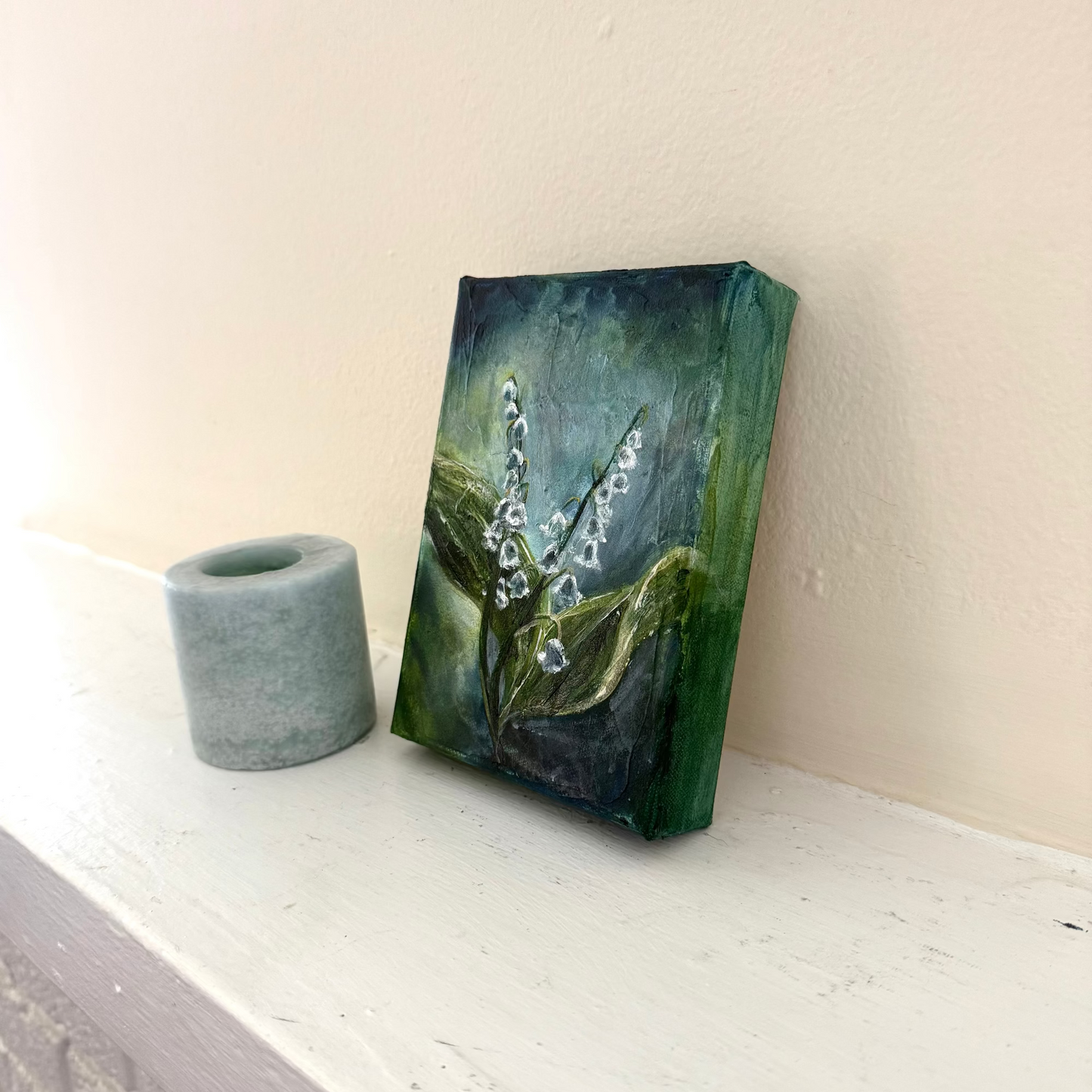 Lily of the Valley flower floral acrylic palette knife painting by Allison Victoria deep greens and blues cool tones and texture on gallery wrapped canvas styled delicately next to a blue candle on a neutral mantle home decor angled shot