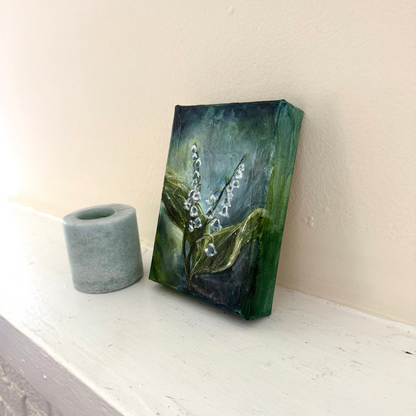 Lily of the Valley flower floral acrylic palette knife painting by Allison Victoria deep greens and blues cool tones and texture on gallery wrapped canvas styled delicately next to a blue candle on a neutral mantle home decor angled shot