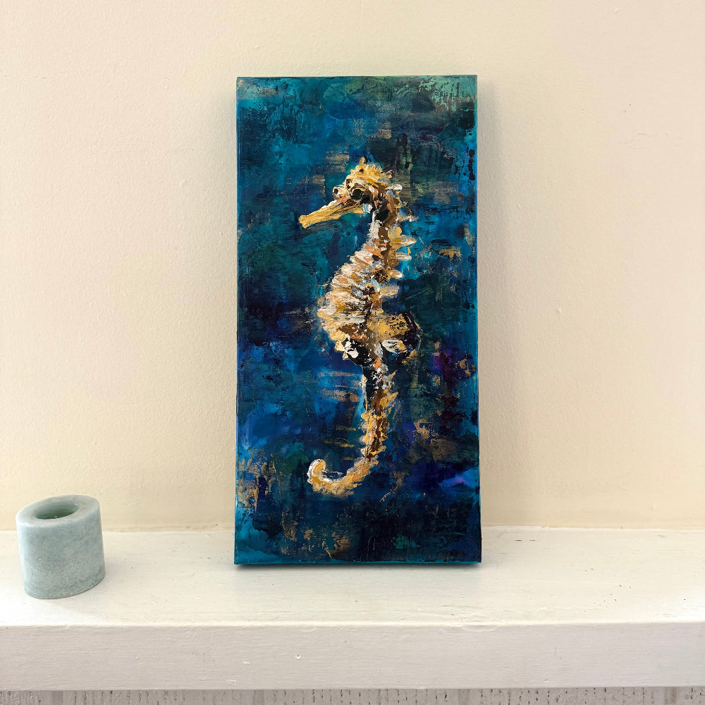 Painting of a seahorse on a blue background with a candle next to it on a neutral light surface on a mantle
