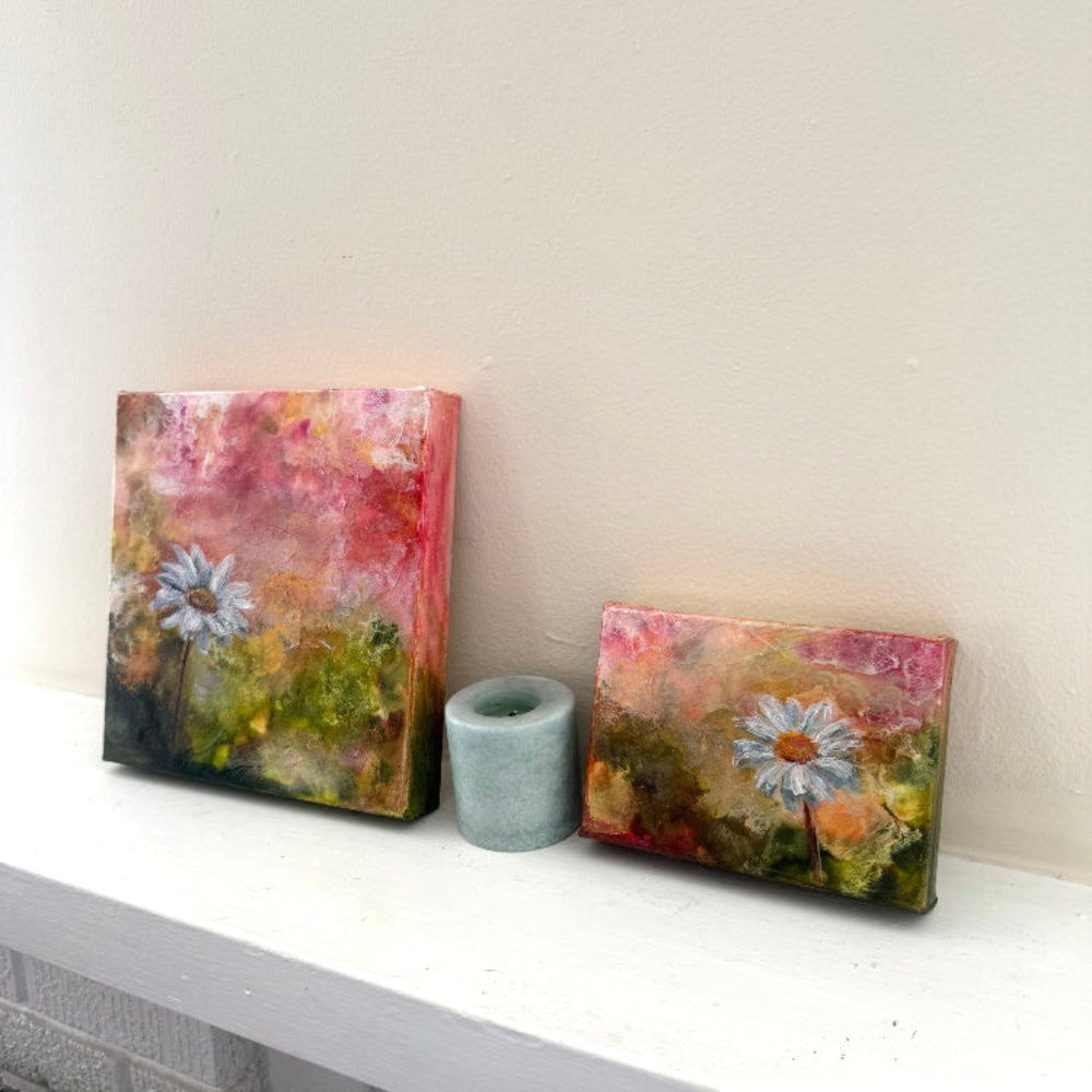 Two small floral paintings on a shelf with a candle between them. Birth month flower s May bloom daisy studies I and II acrylic palette knife paintings on neutral mantle arranged decor